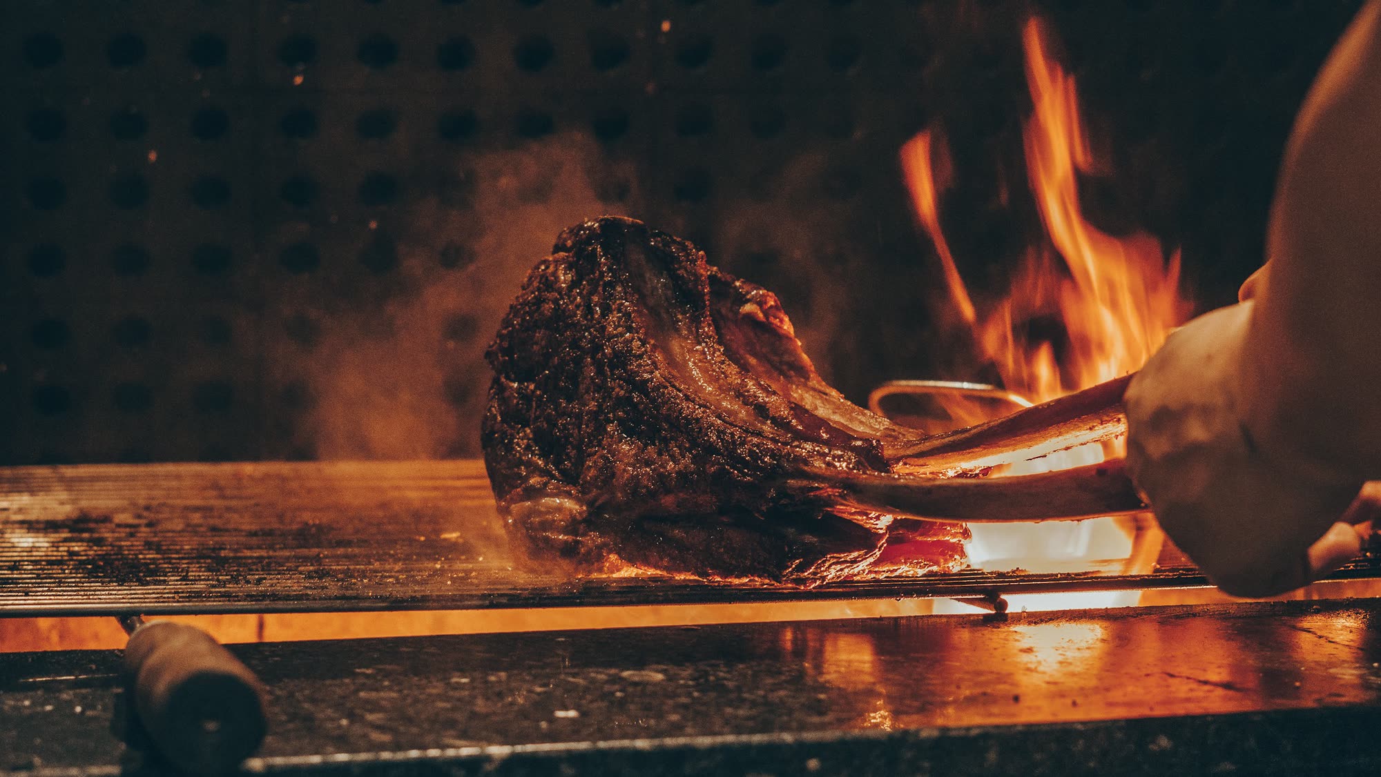 A tomahawk steak on the grill with oak flames behind it